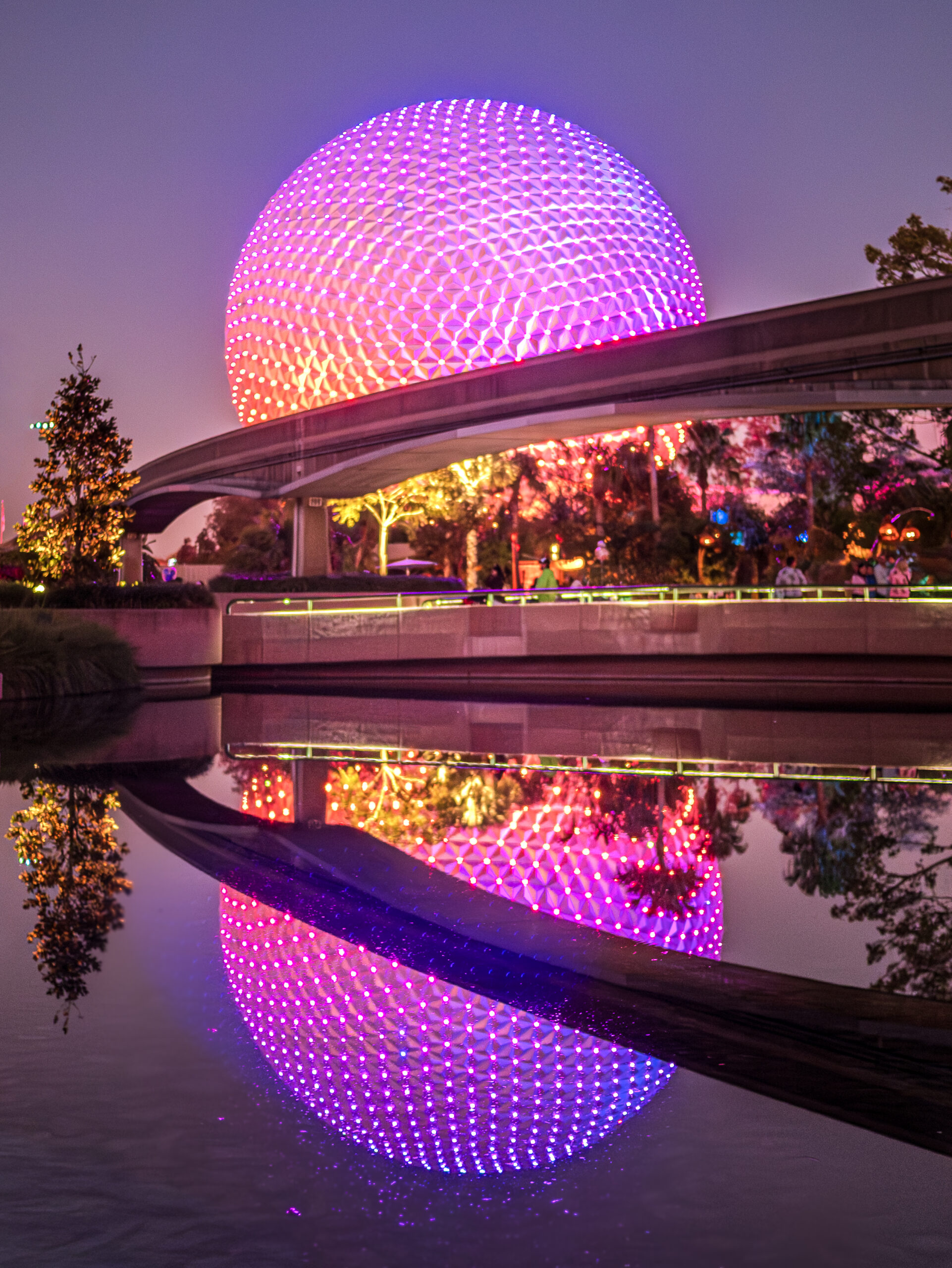 Spaceship Earth at EPCOT glowing in purple and pink LED lights at night, with its full reflection mirrored in the still lagoon below.