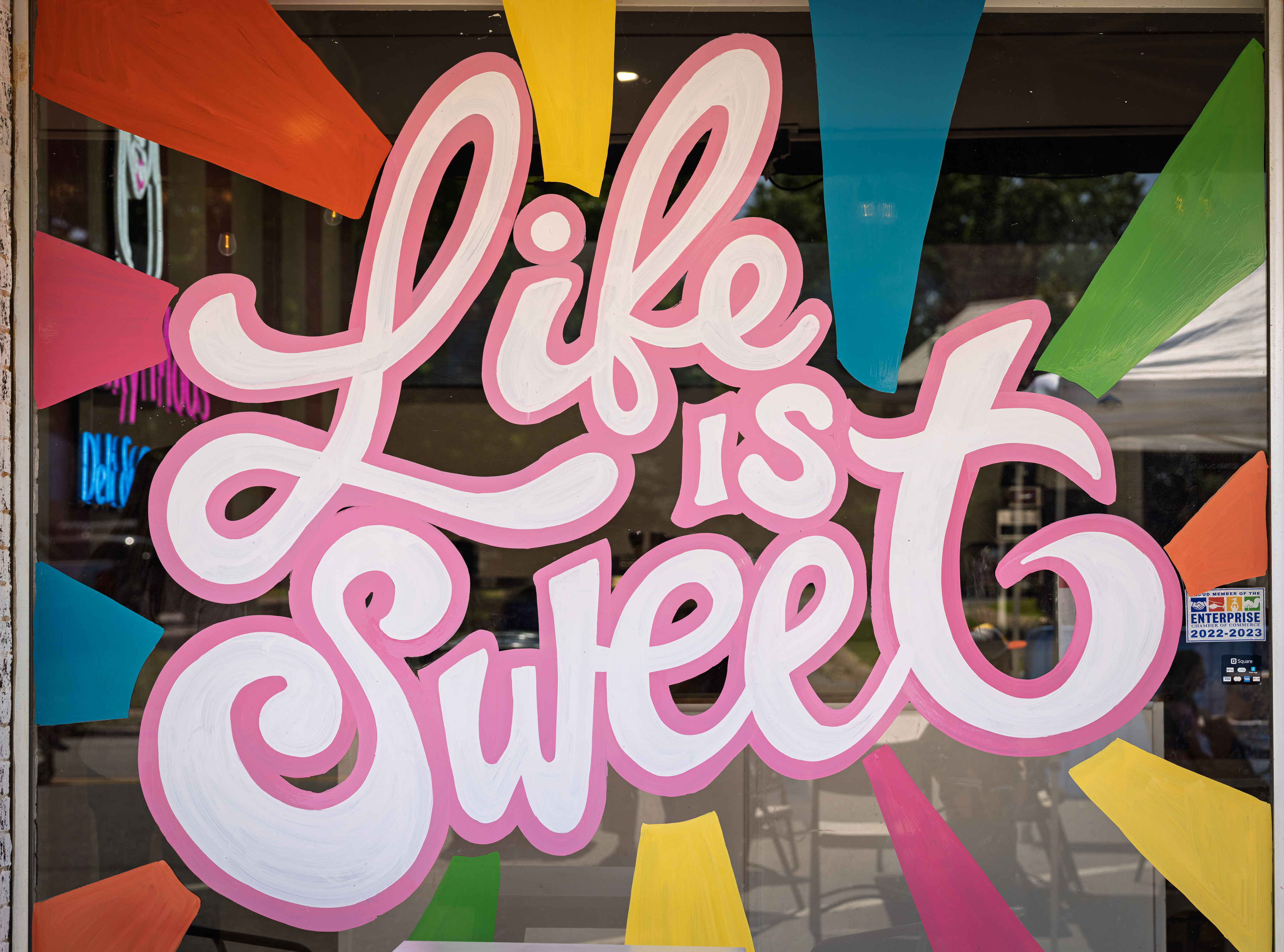Hand-painted shop window mural with the words “Life is Sweet” in pink and white script, surrounded by colorful geometric bursts.