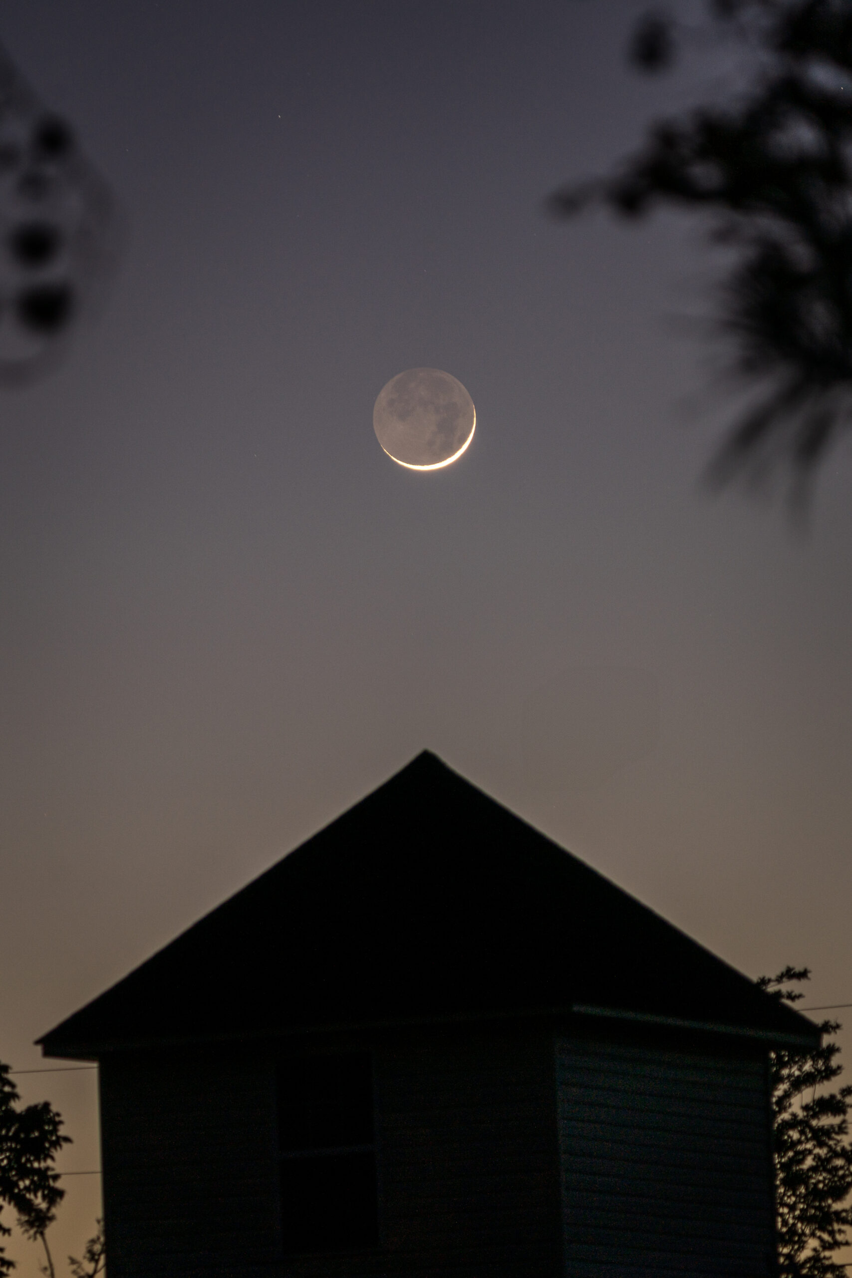 Crescent moon with visible earthshine above the silhouette of a house roof at dusk, framed by faint tree branches and a gradient evening sky